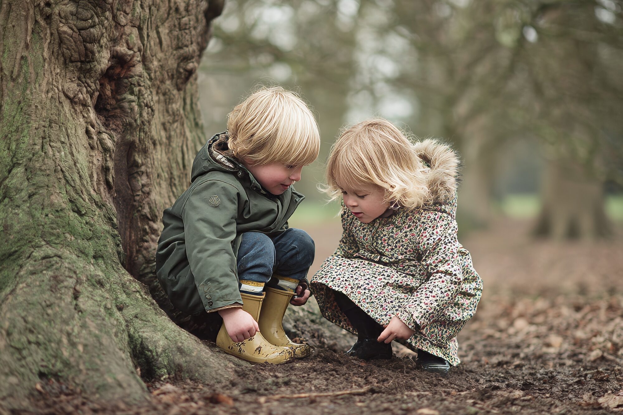 Two young children are engaged in exploration, crouching beside a large tree in a tranquil woodland area on a chilly day paleo chair