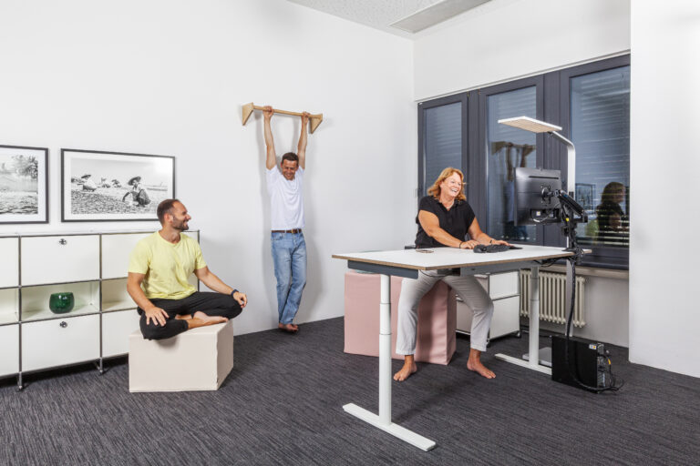 Büro mit höhenverstellbaren Schreibtisch, 2 Personen sitzen auf paleo chairs, ein Mann hängt an der paleo bar. Alle 3 lachen.